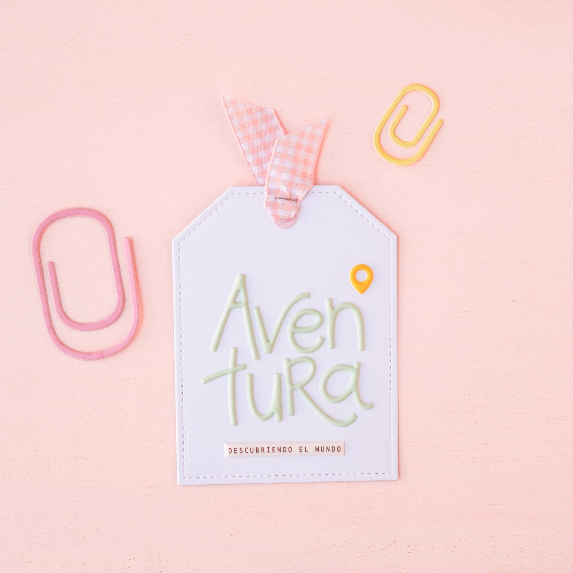 Etiqueta branca em formato de tag com laço xadrez rosa e branco e texto colorido rodeada por clips coloridos sobre fundo rosa claro.