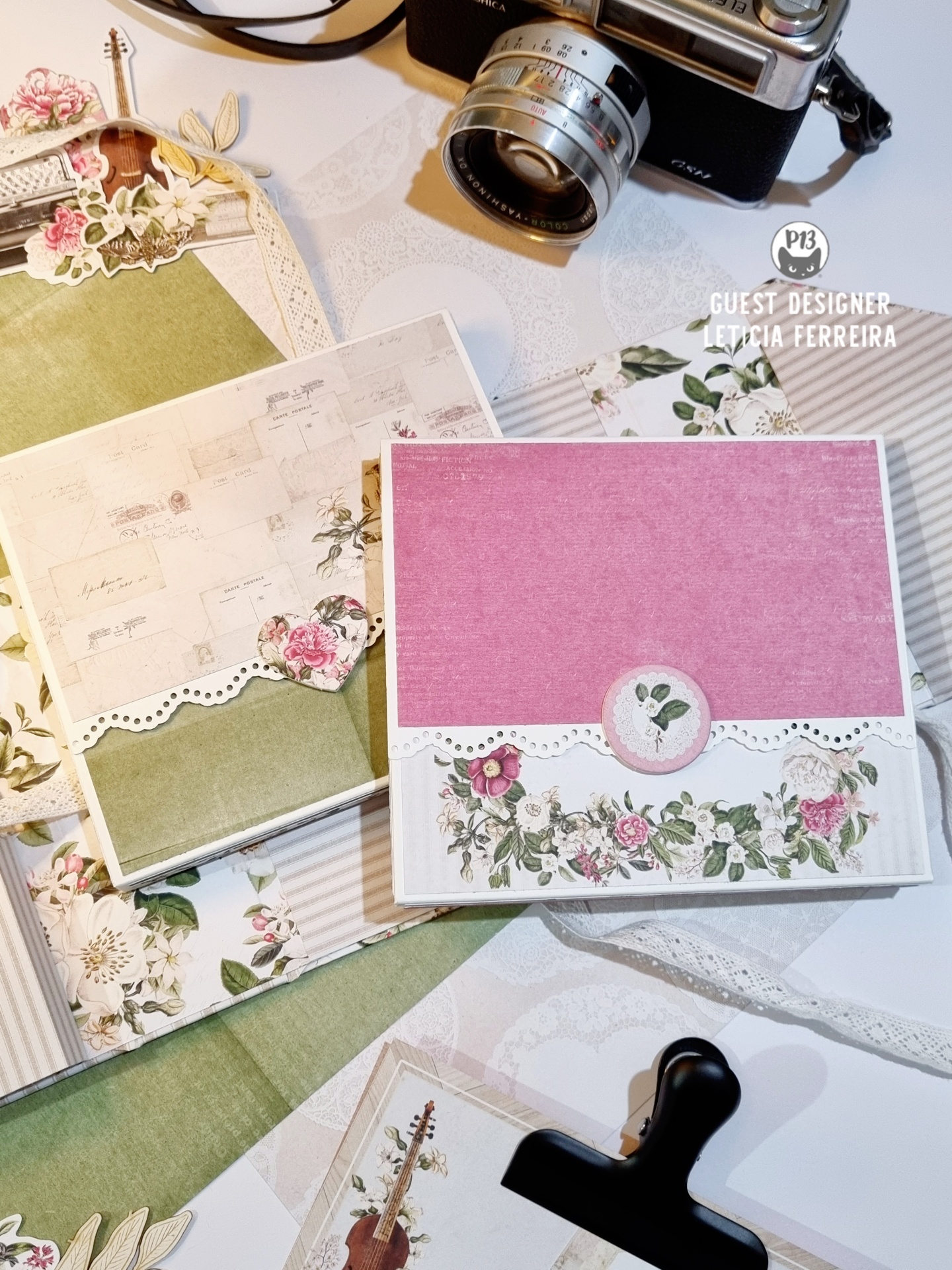 Dois álbuns de scrapbook com capas florais e detalhes rendados sobre mesa branca com câmera fotográfica e clip.