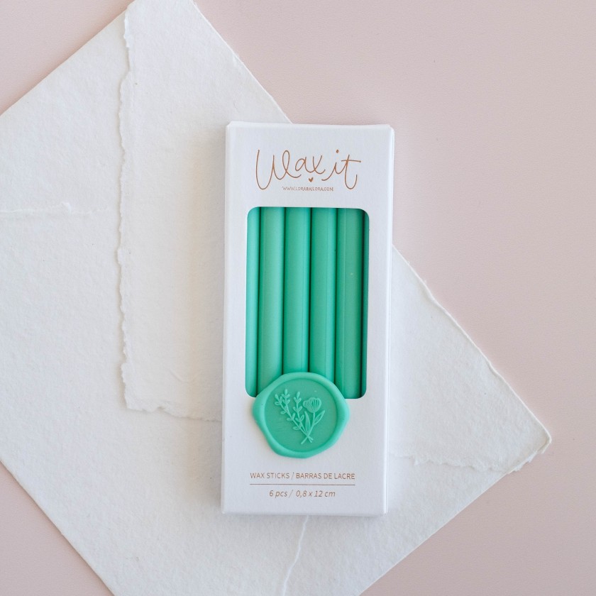 Caixa branca com varetas de cera verdes para vedar e selo decorativo verde, sobre folhas de papel branco e rosa