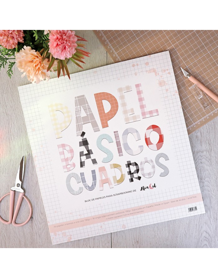 Bloco de papel para scrapbooking com letras coloridas e quadriculadas, tesoura e flores ao fundo.