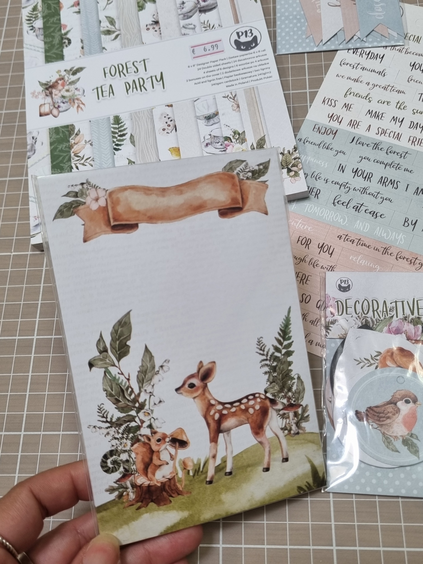 Papel decorativo com desenho de veados e esquilo em floresta, embalado em plástico, com blocos de papel e etiquetas ao fundo.