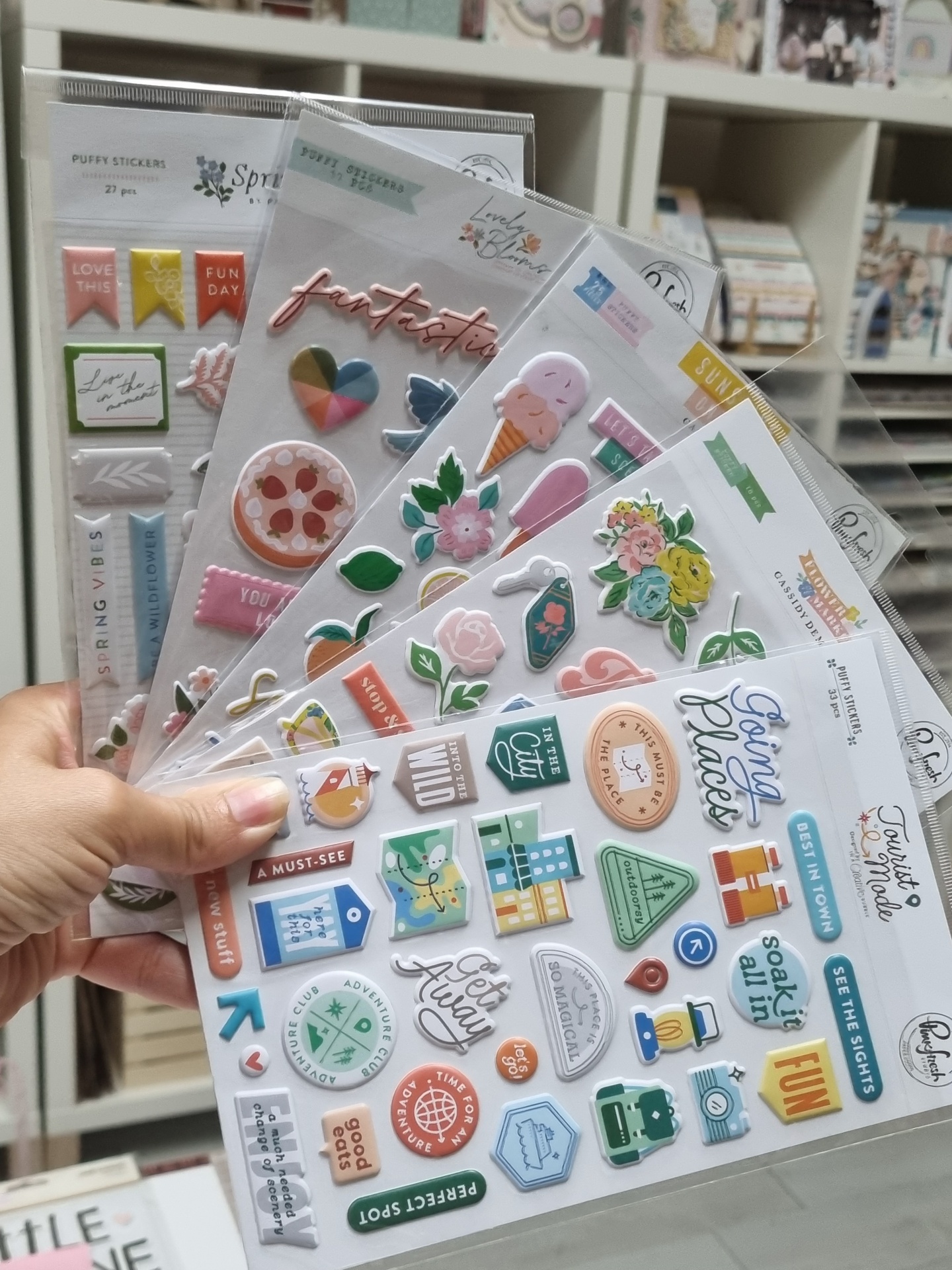 Folhas de autocolantes 3D coloridos com padrões de flores, comida e textos, seguradas por uma mão num ambiente com prateleiras no fundo.