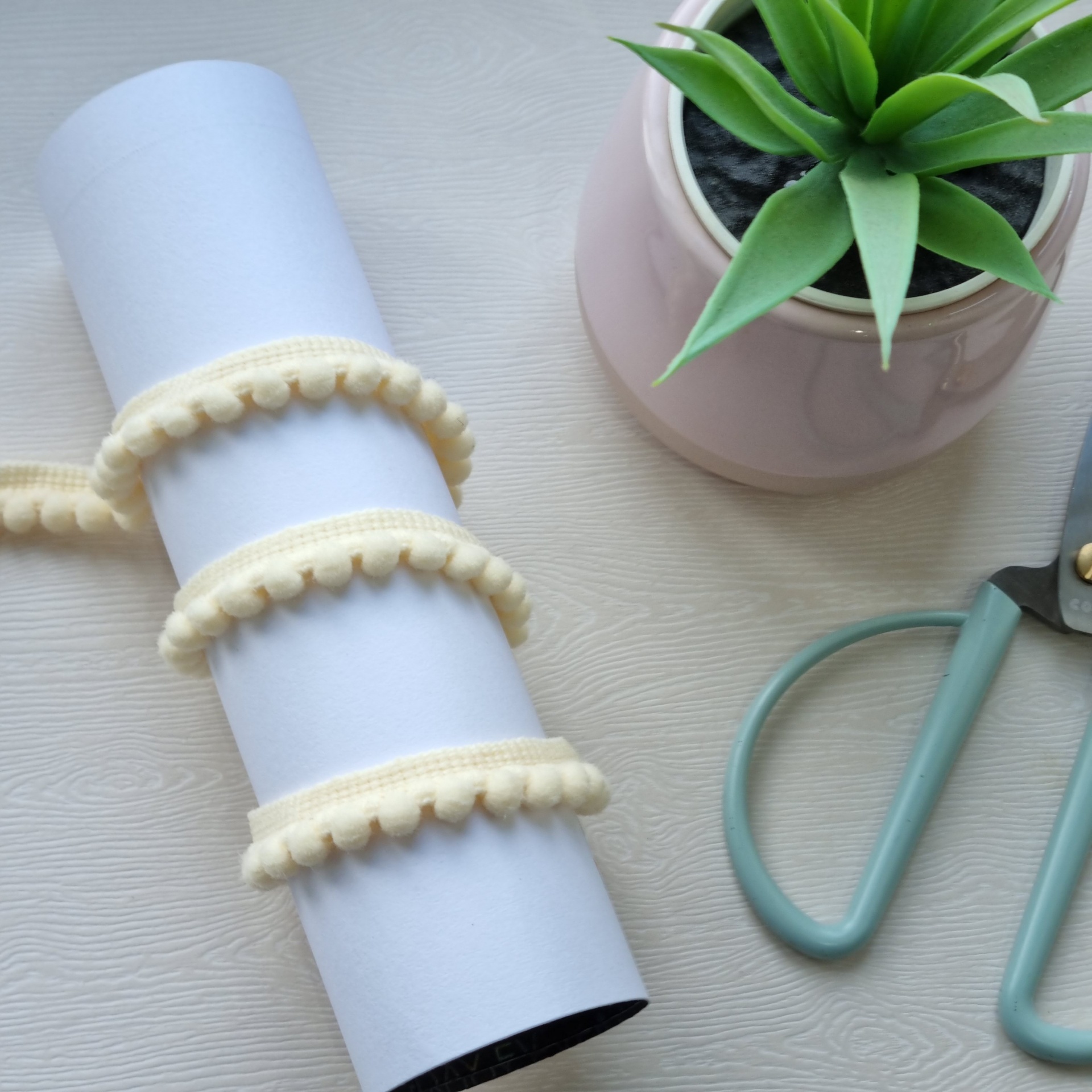 Rolo branco com fita branca de pompons, planta em vaso cor de rosa e tesoura com cabo cinza