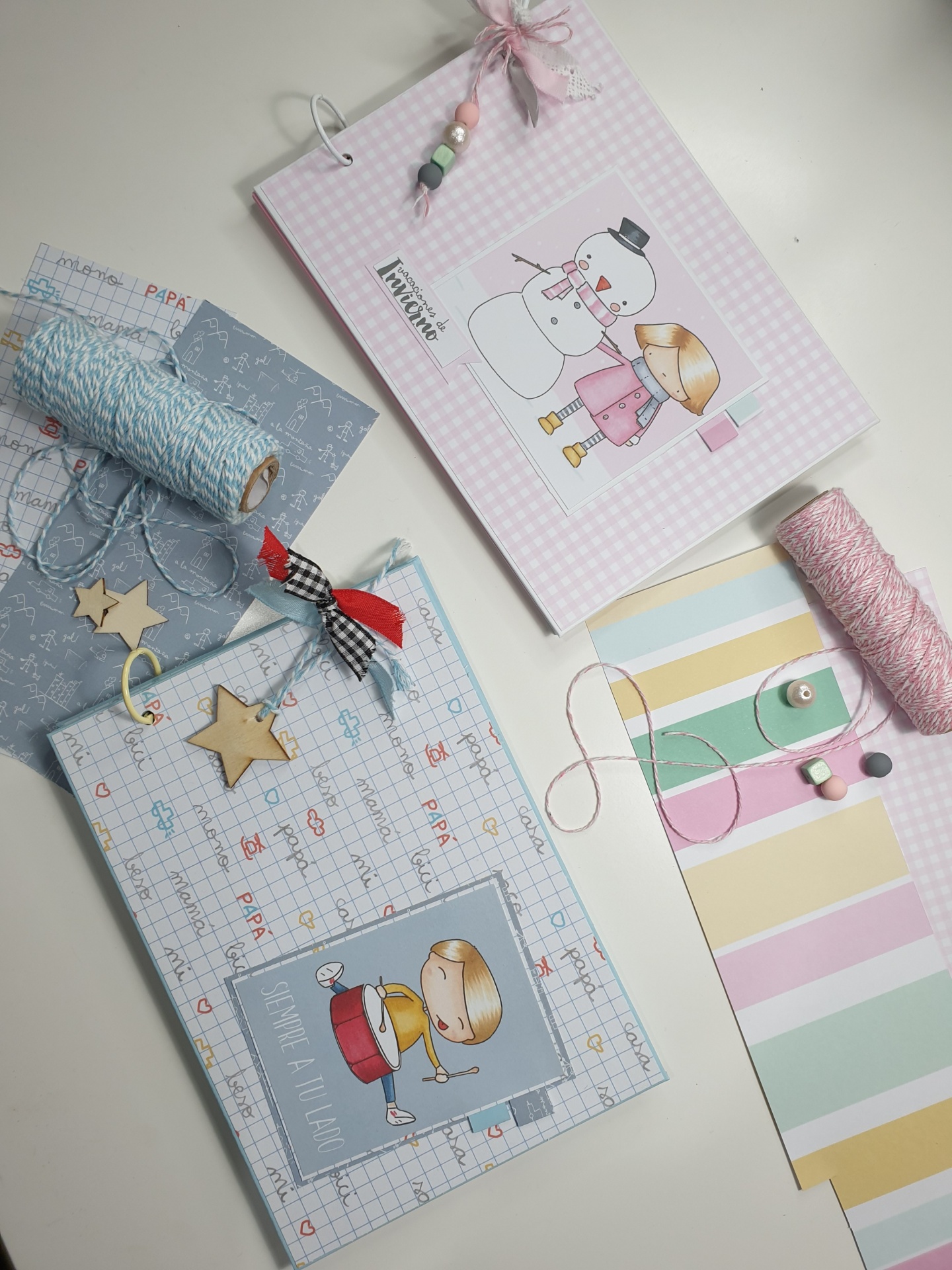 Dois álbuns infantis decorados com desenhos de boneco de neve e menina, e menina loira, sobre mesa com rolos de fio e papel decorativo
