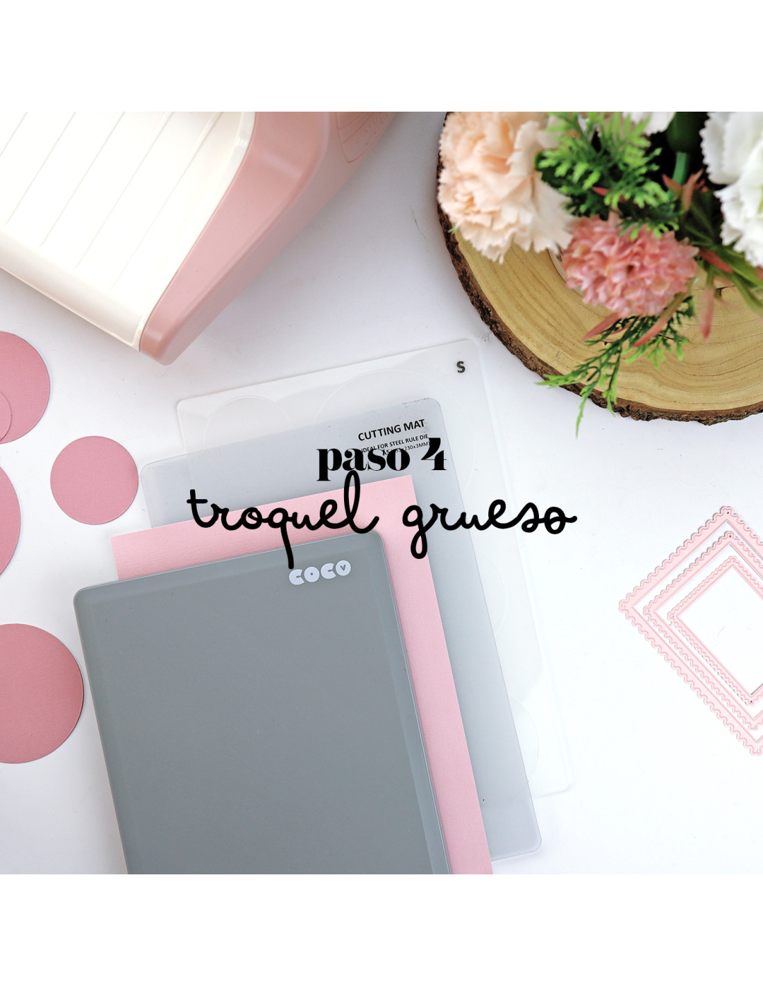 Acessórios de artesanato para corte com tapetes e recortes em tons rosa, cinzento e transparente em superfície branca ao lado de flores