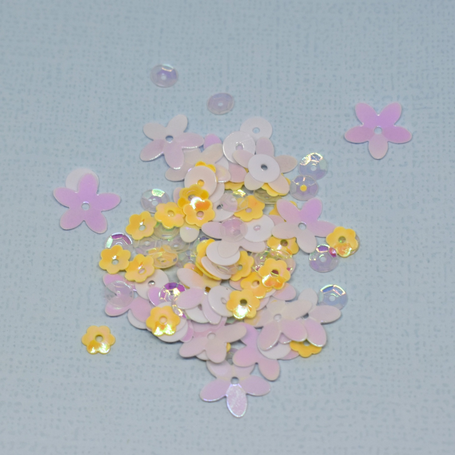 Lantejoulas em forma de flor amarelas, rosa e transparentes sobre fundo cinzento claro