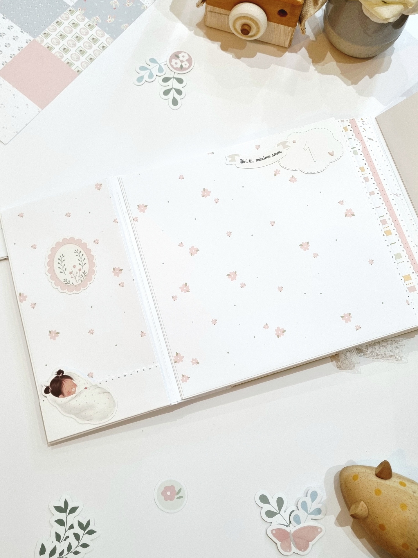 Livro scrapbook aberto com padrão floral e decoração de bebé e autocolantes sobre mesa branca.