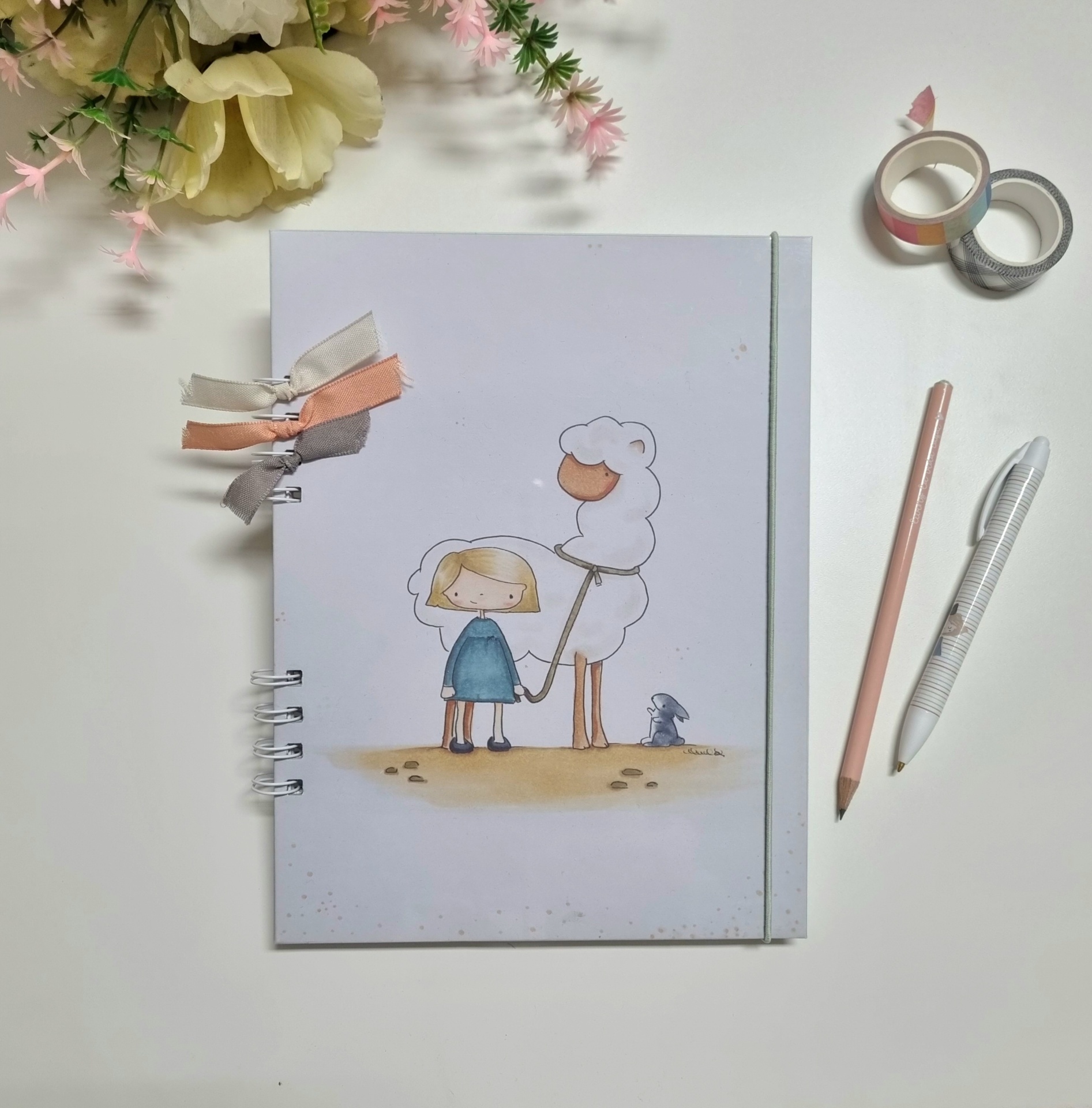 Caderno ilustrado com menina, ovelha e rato e acessórios de escrita ao lado