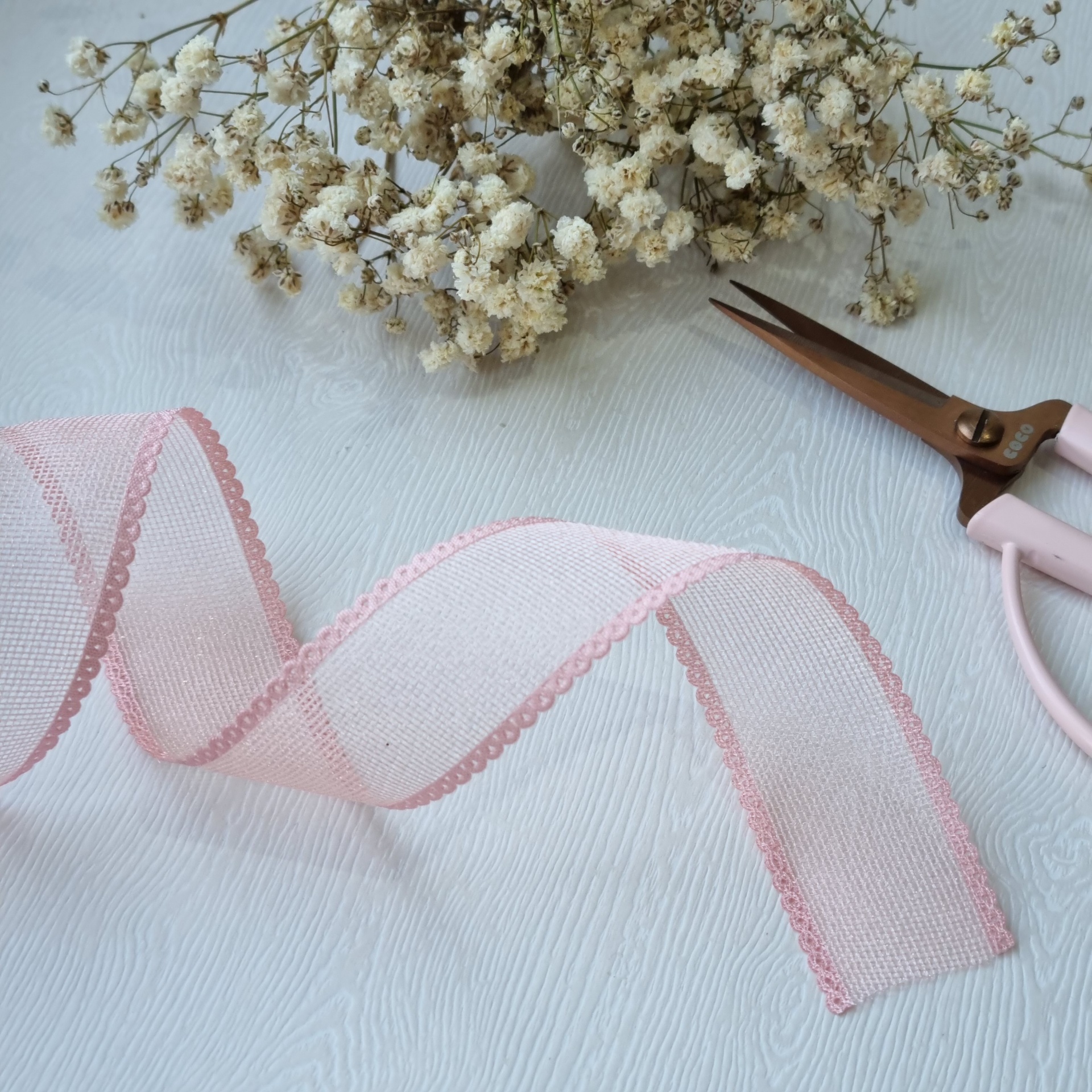 Fita decorativa rosa transparente com bordas onduladas ao lado de flores secas e tesoura rosa