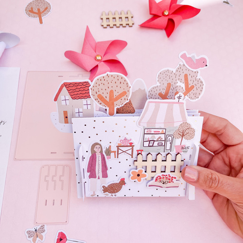 Produto artesanal em papel ilustrado com casa, árvores, mulher e mercado sobre mesa rosa.
