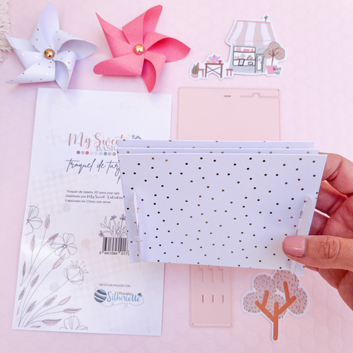 Folha de papel branco com bolinhas douradas segurada sobre fundo rosa com decoração de papéis e pinwheels