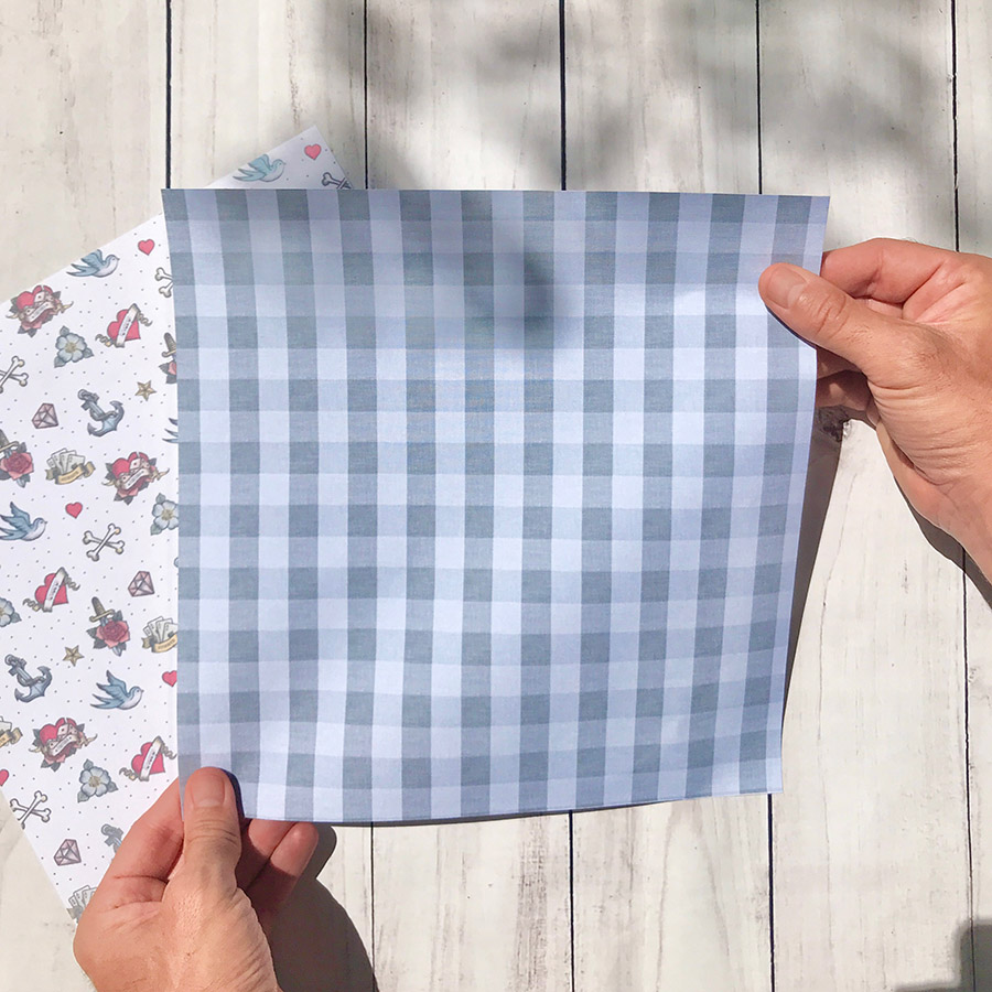 Dois lenços quadrados com padrões diferentes, um xadrez azul e branco e outro com desenhos coloridos, segurados em mãos contra madeira clara.