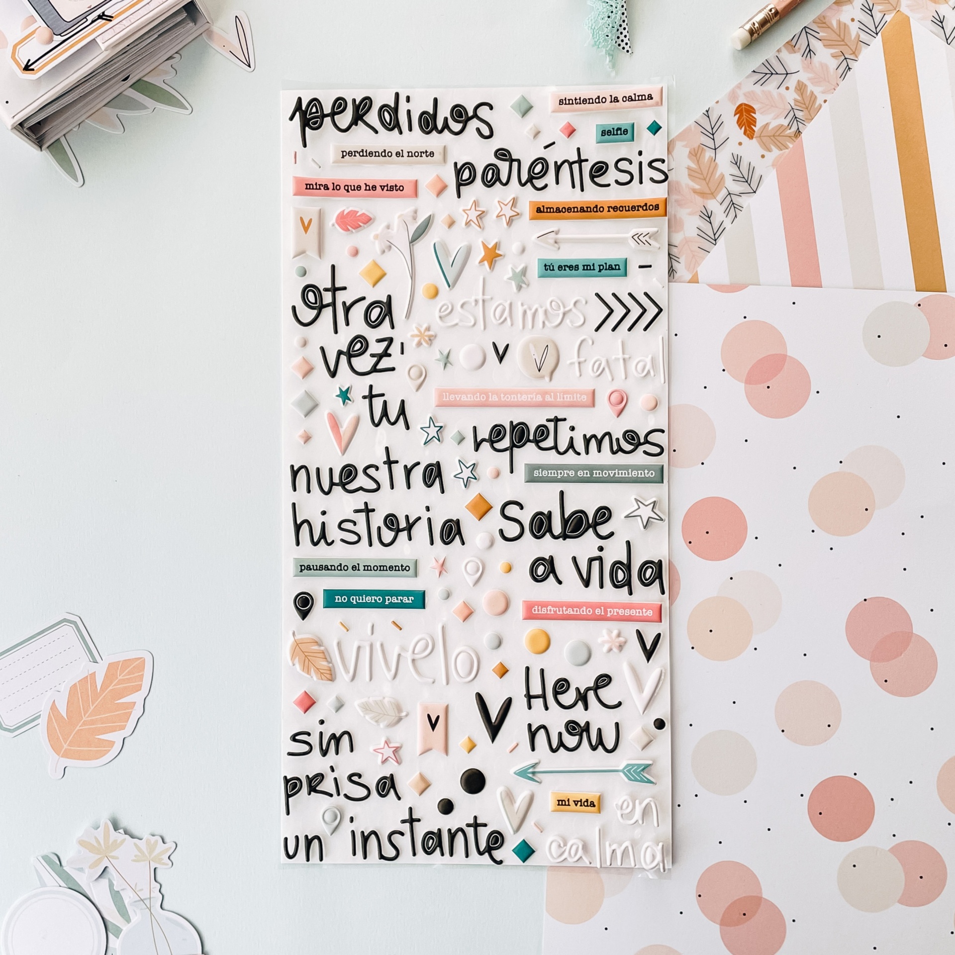 Folha translúcida com adesivos de palavras e frases em espanhol sobre mesa clara com folhas decoradas em tons pastel