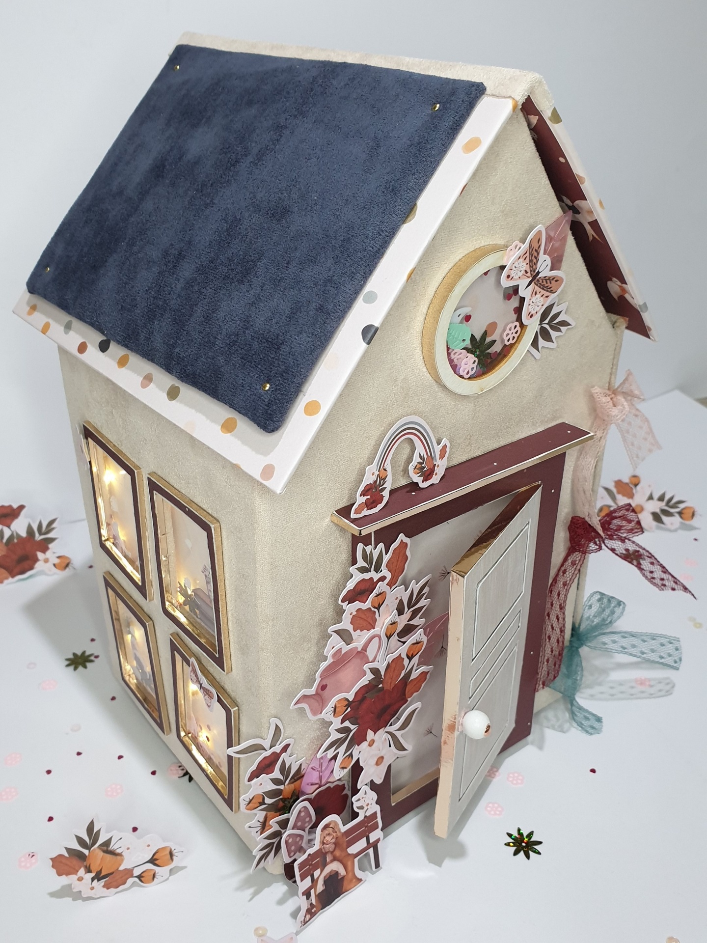 Casa decorativa em tecido cinzento claro com telhado azul e janelas iluminadas