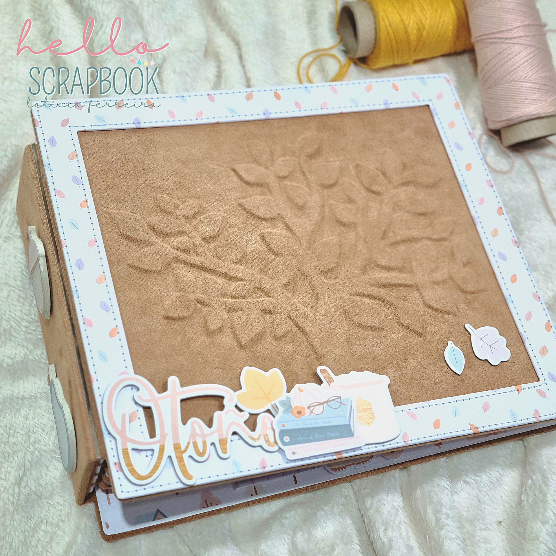 Álbum scrapbook com capa texturizada e decoração em papel, cones de fios ao fundo