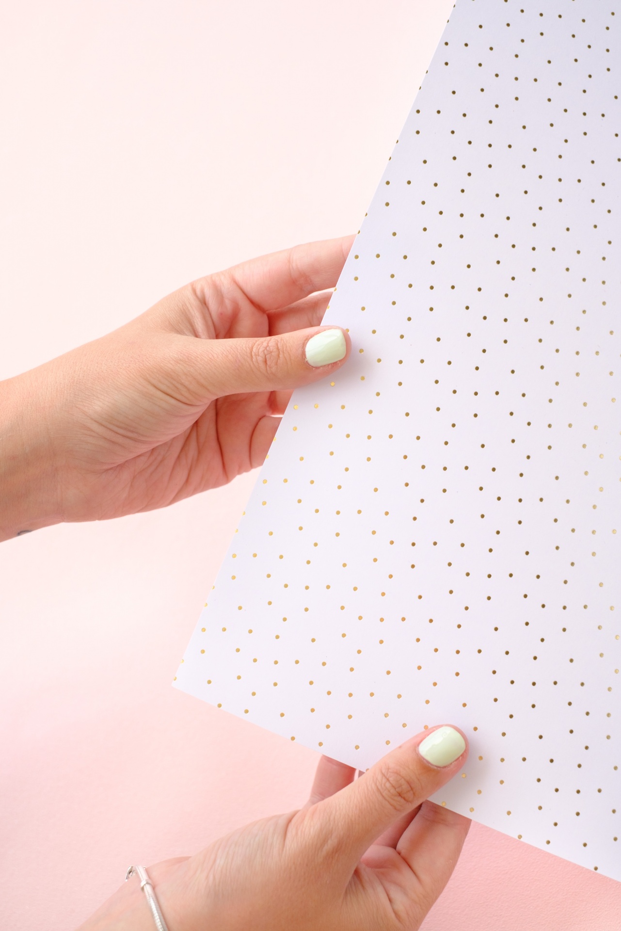 Folha branca com pontos dourados segurada por mãos com unhas claras, fundo rosa