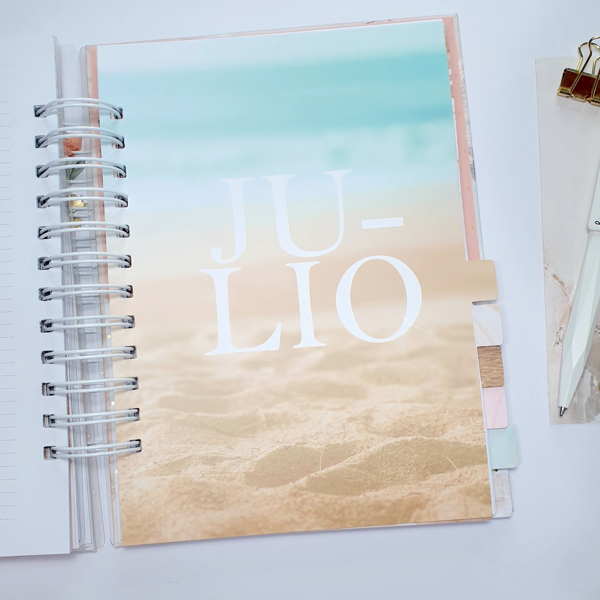 Caderno com capa de praia e texto JULIO, espiral e divisórias coloridas