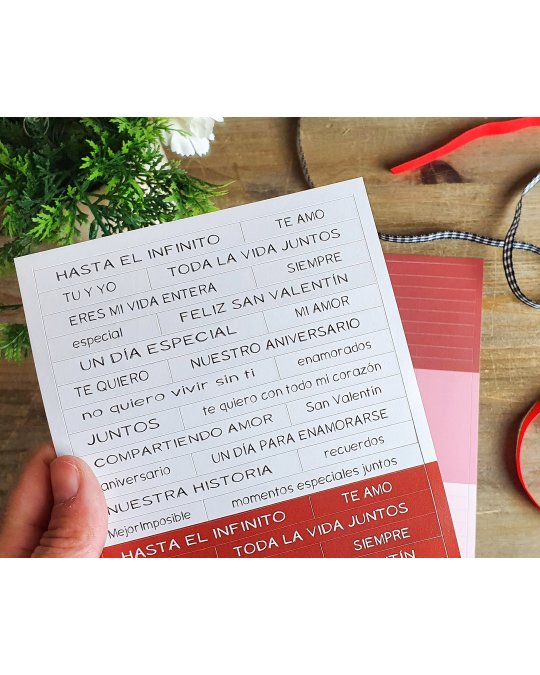 Folha com frases em castelhano para cartão, mão segurando, fundo de madeira, plantas e fitas ao redor