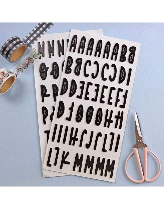 Folhas com letras autocolantes negras, rolos de fita adesiva decorativa de bicicletas e xadrez, e tesoura rosa em fundo azul.