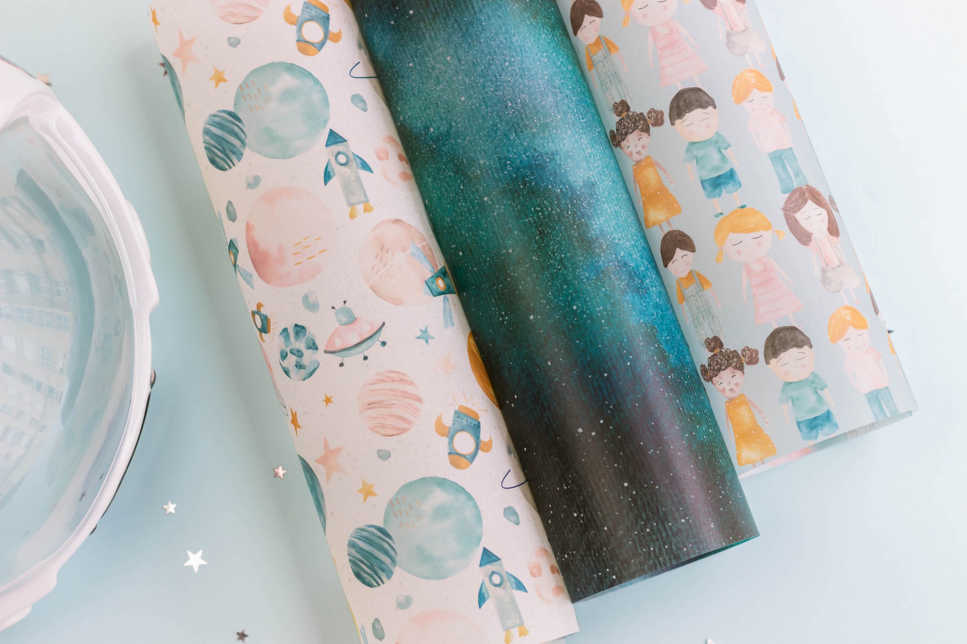 Três rolos de papel decorativo com temas espaciais e crianças em estilo cartoon sobre fundo claro