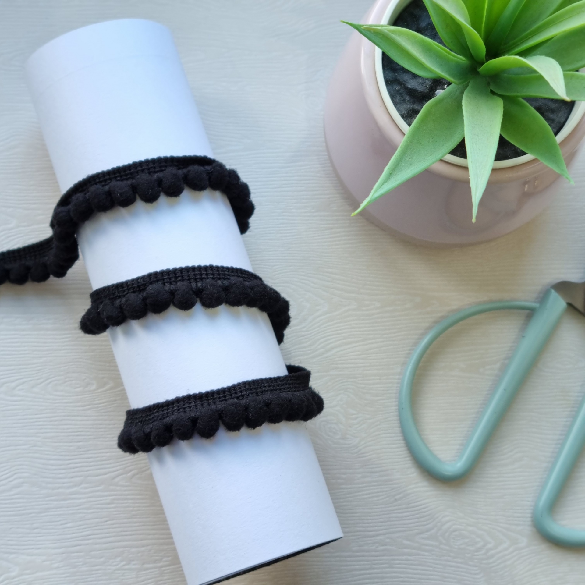 Fita preta com pompons enrolada num rolo branco ao lado de planta e tesoura.