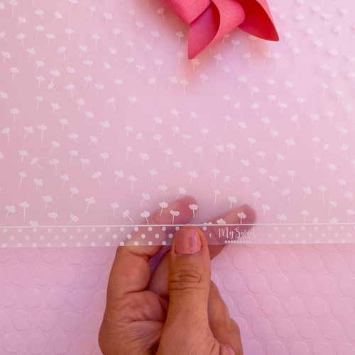 Mão segurando folha translúcida com desenhos de dentes-de-leão e selo 'My Sweet' sobre fundo rosa texturizado e laço rosa