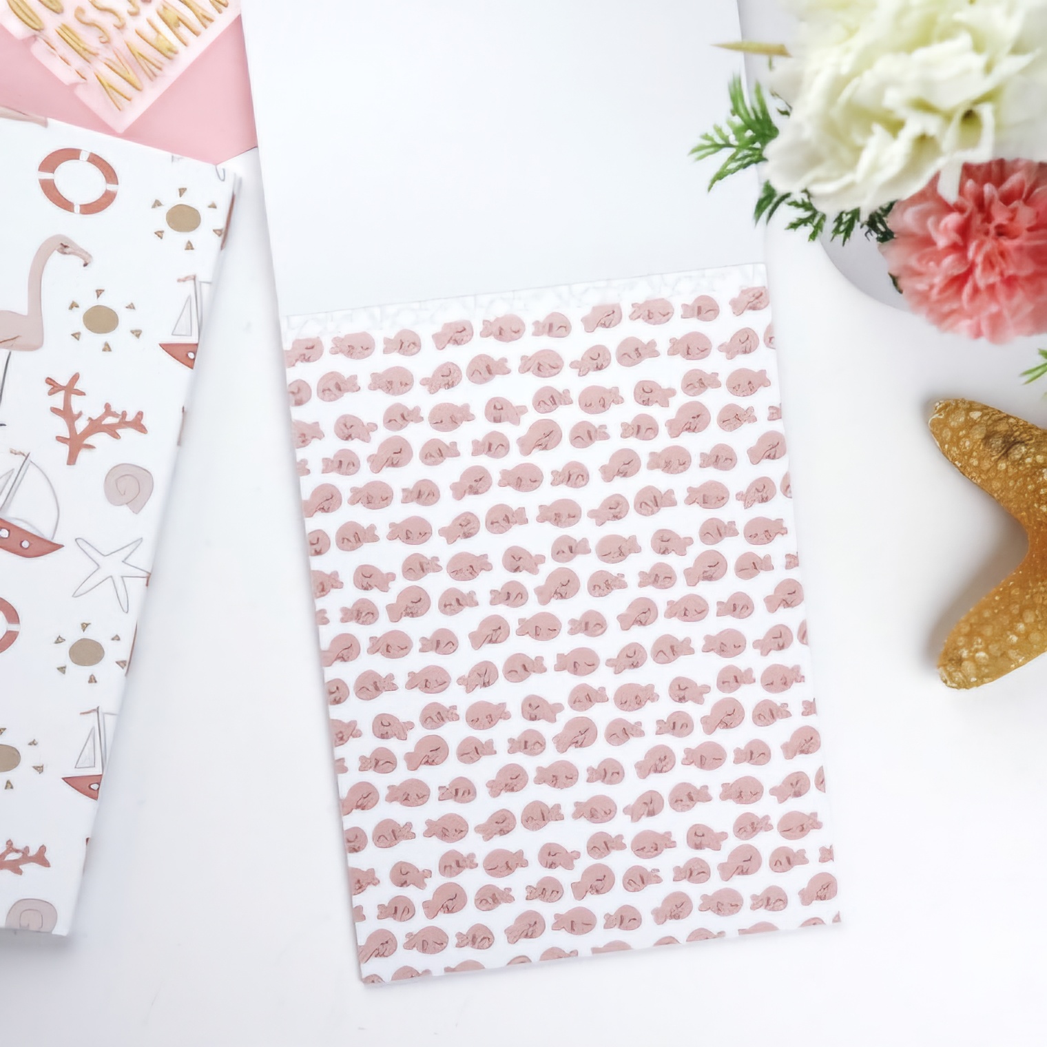 Bloco de papel com padrão de conchas rosas sobre mesa branca com livro infantil e decoração de flores e estrela do mar
