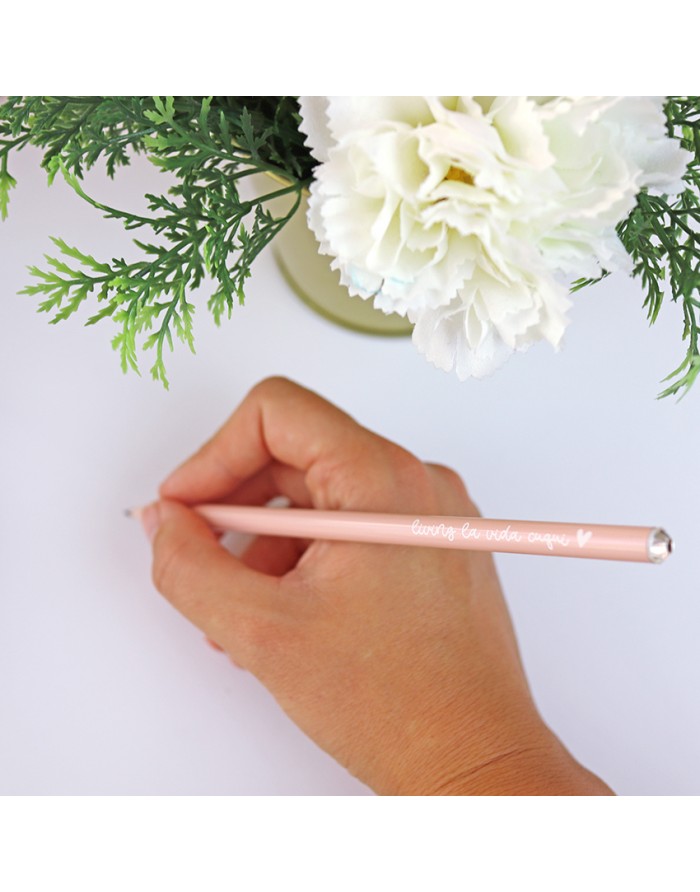 Mão segurando caneta cor-de-rosa com texto branco escrevendo num papel branco perto de flores