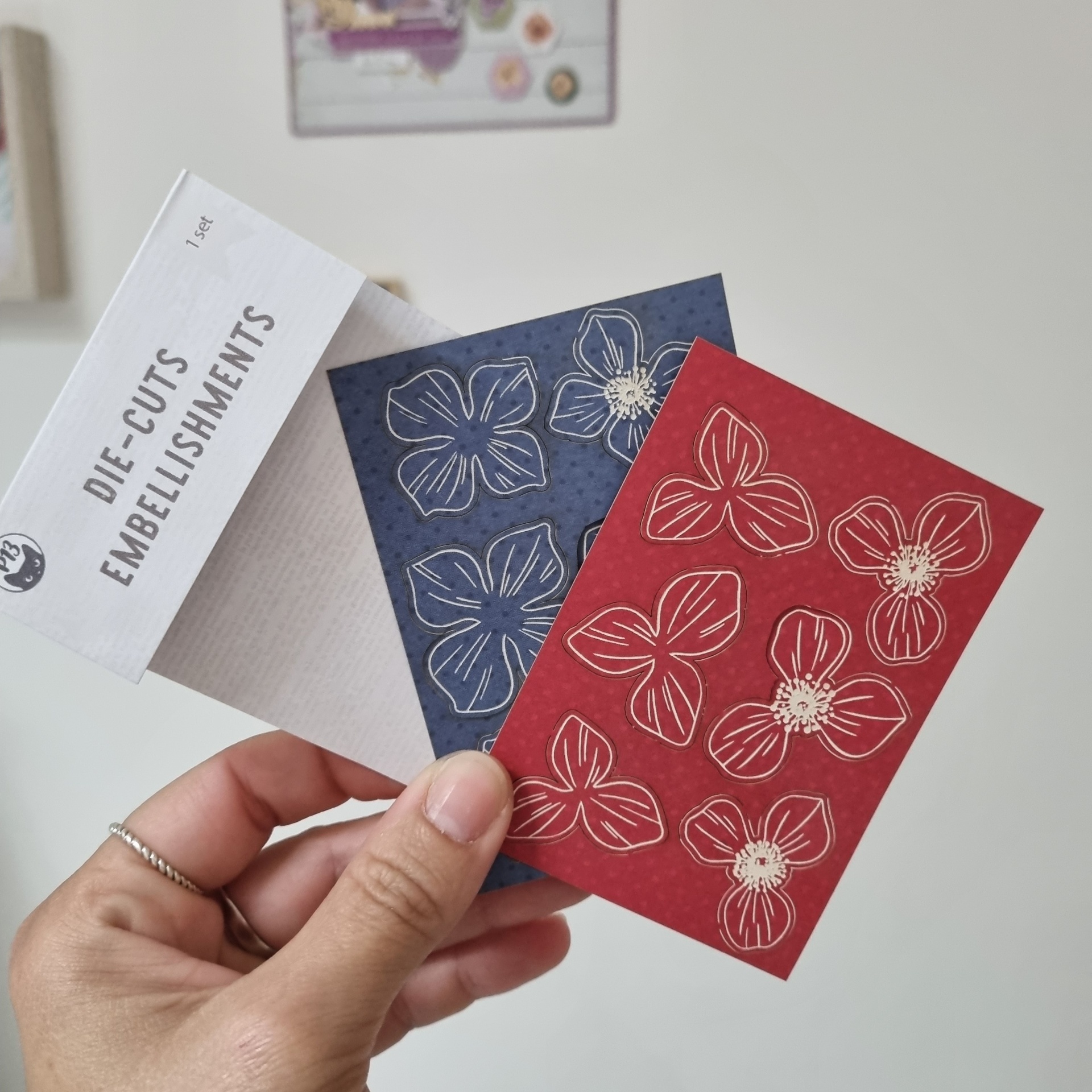 embalagens de papel azul e vermelha com padrão floral seguradas por mão e embalagem branca com texto DIE-CUTS EMBELLISHMENTS