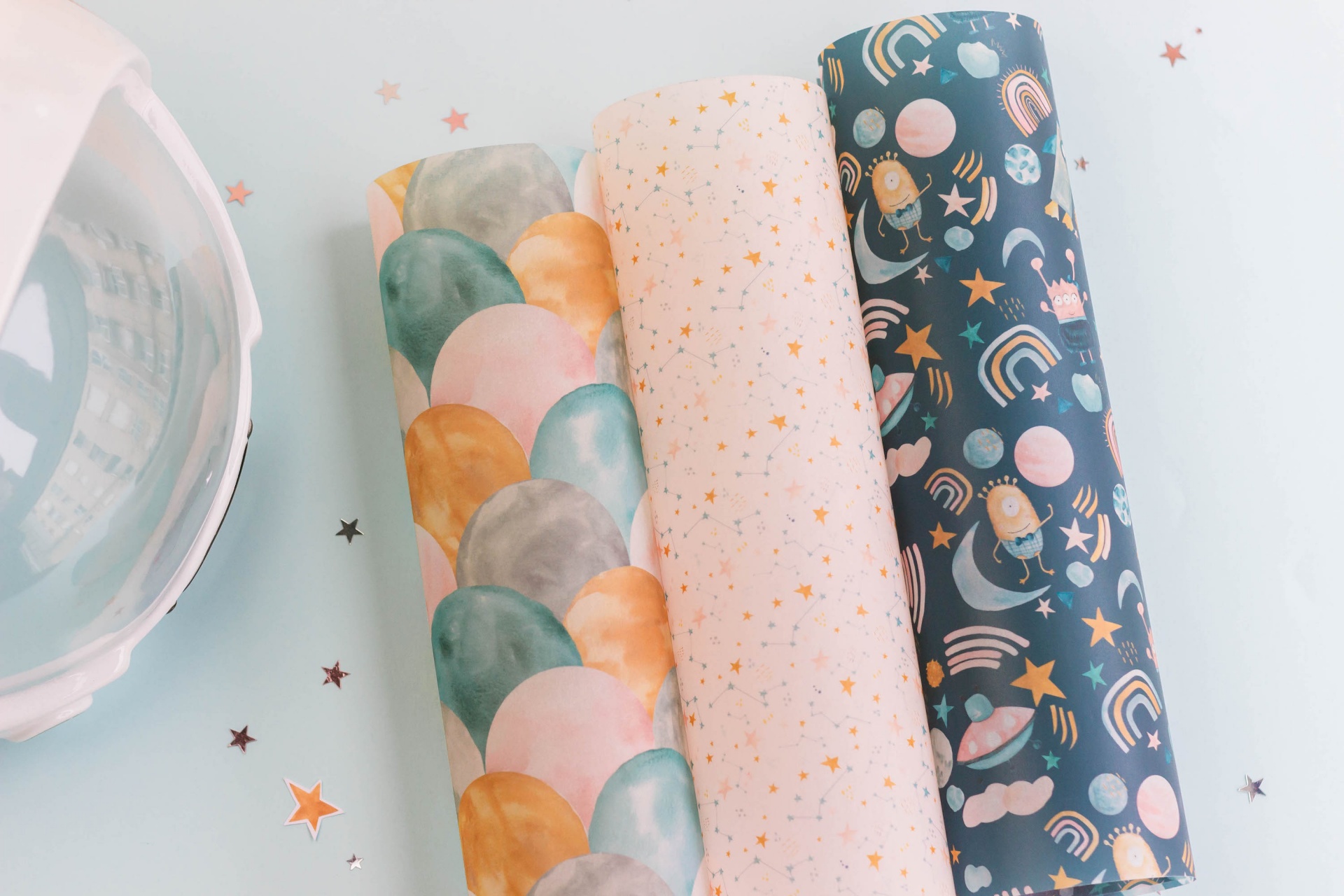 Três rolos de papel colorido com padrões variados em cima de uma superfície clara ao lado de um prato branco