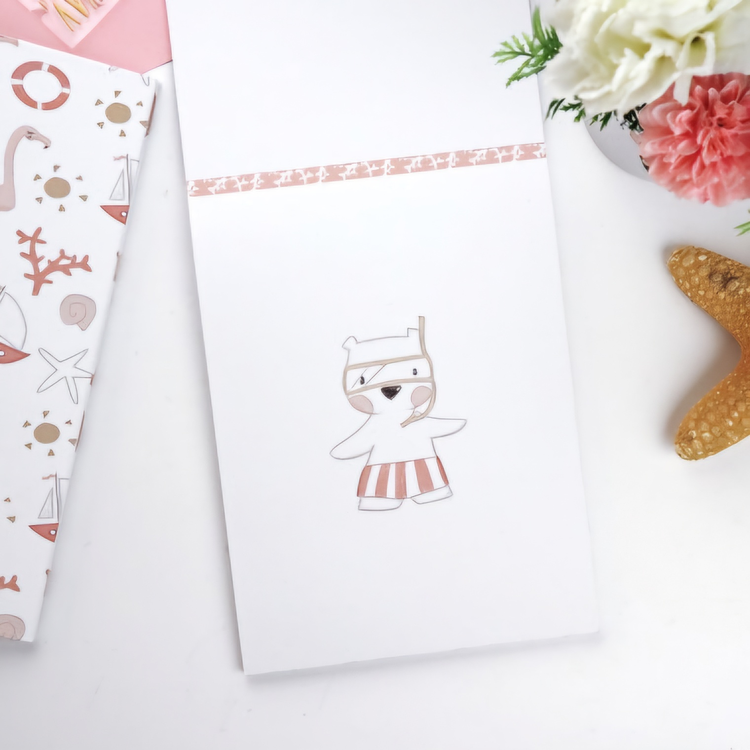 Caderno branco com desenho de urso rosa e bege, caixa decorada com motivos marítimos e flores numa superfície branca