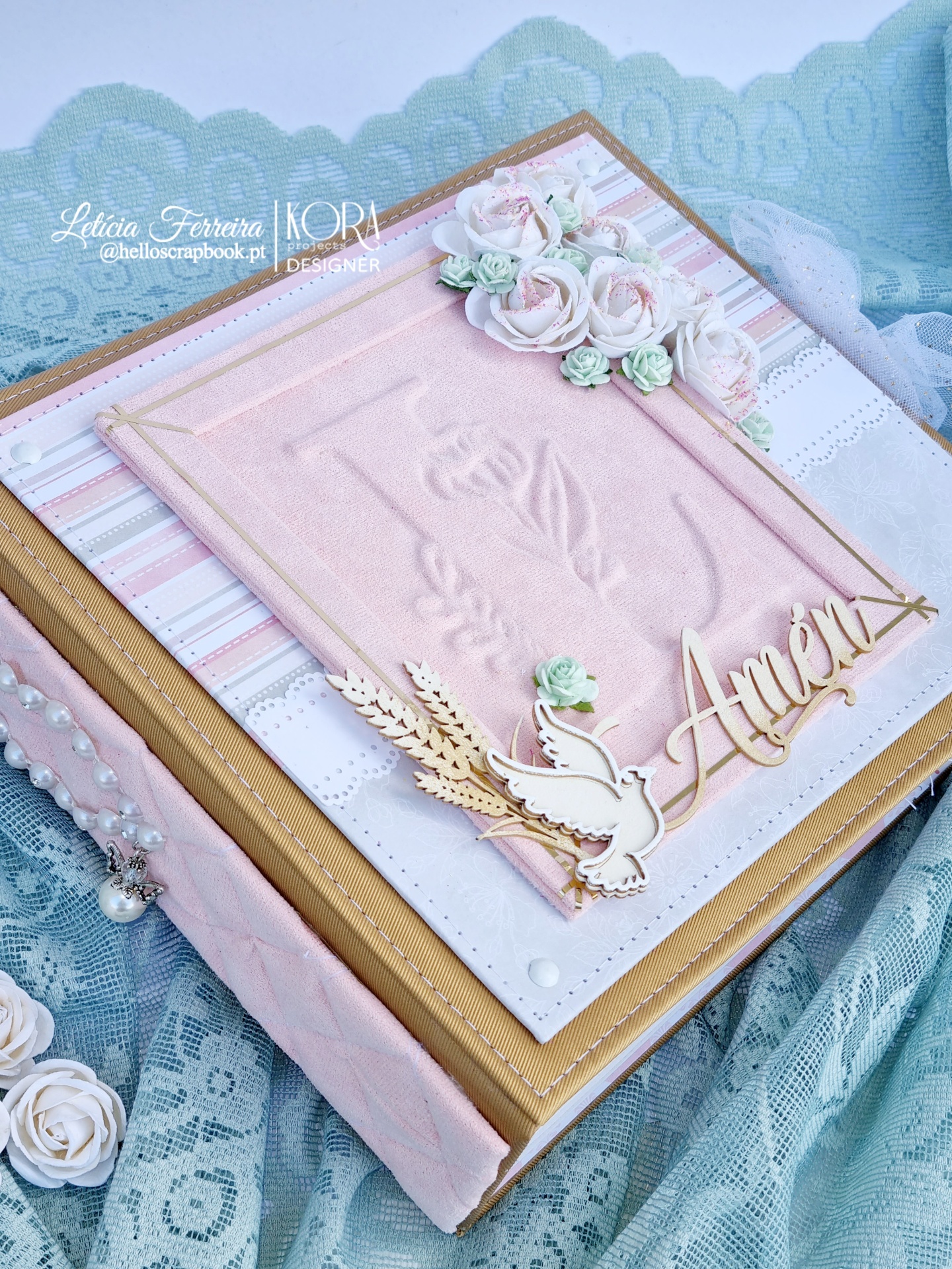 Livro decorativo cor-de-rosa com relevo e adornos florais e em madeira com palavra Amém sobre fundo rendado azul.
