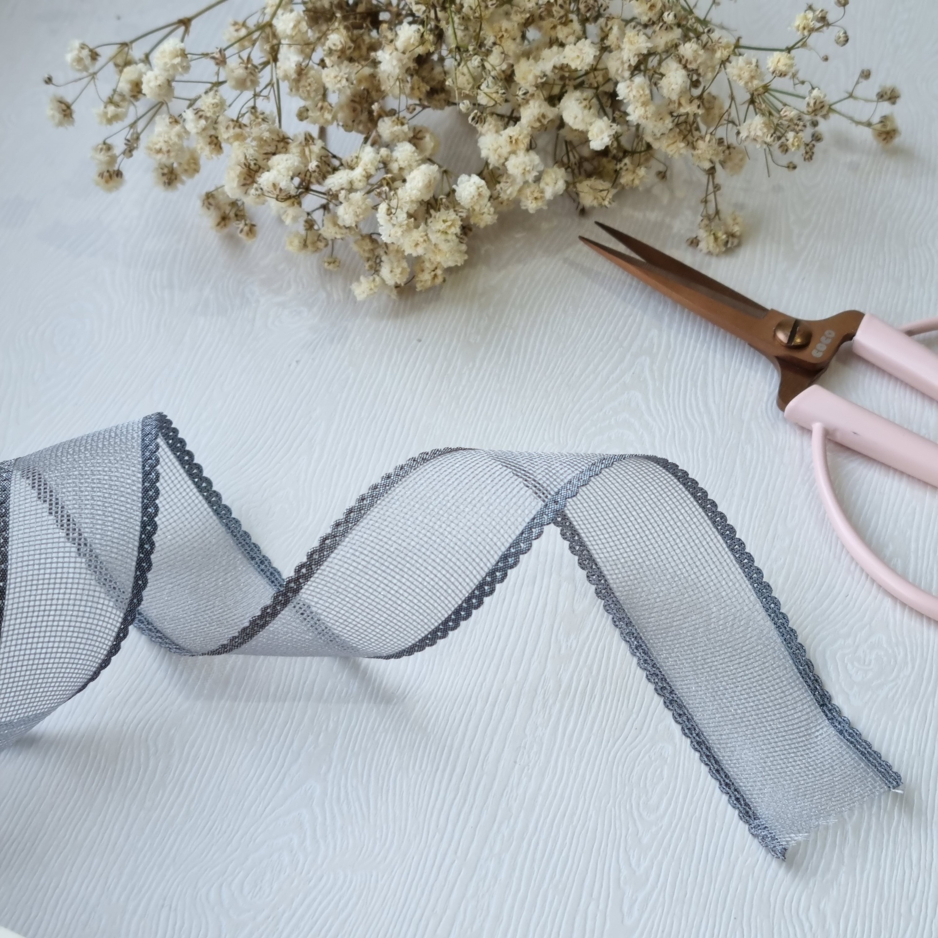 Fita cinza decorativa em organza com bordas bordadas, flores secas e tesoura rosa numa superfície branca.