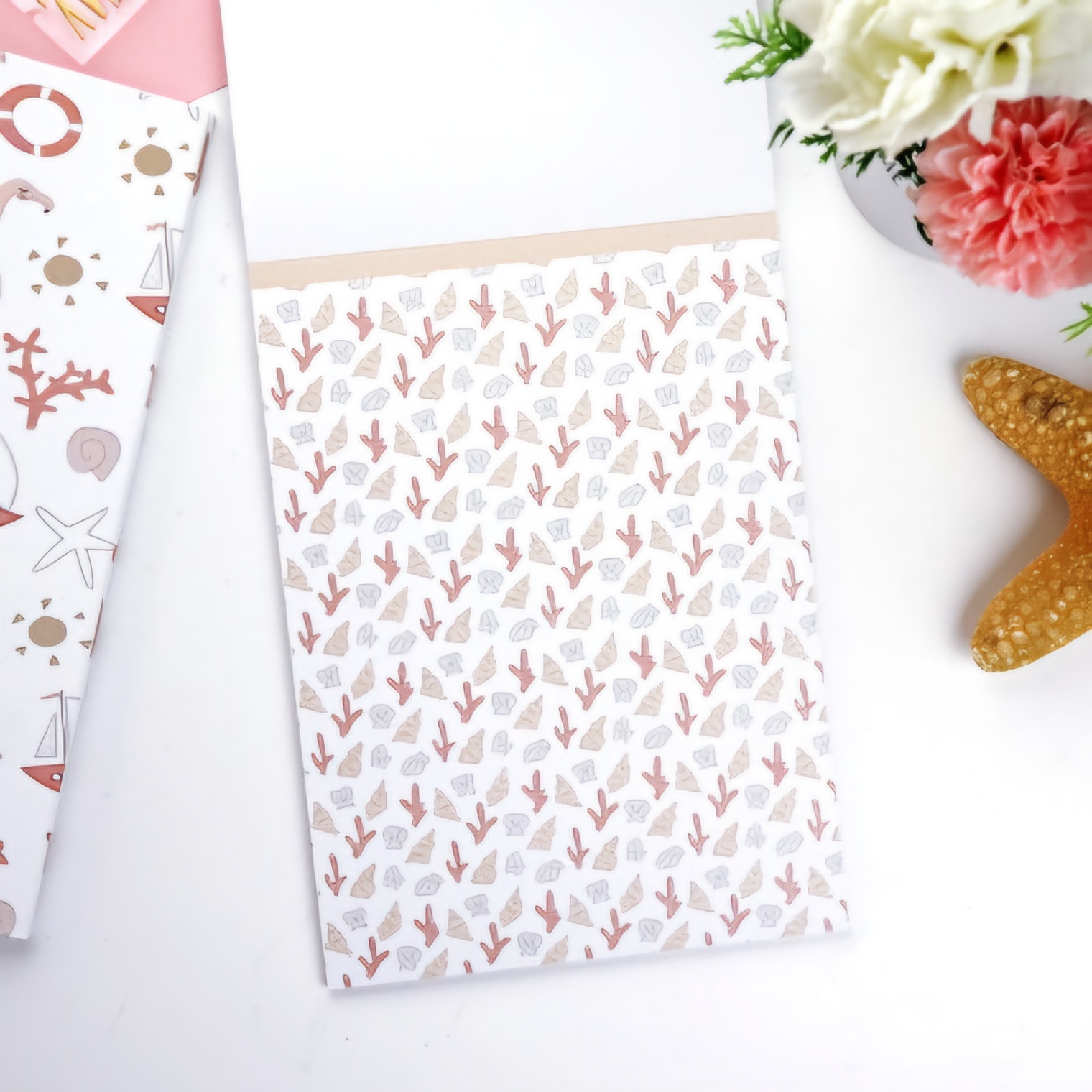 Caderno com padrão de corais em tons castanho, rosa e bege sobre mesa branca com estrelas-do-mar e flores