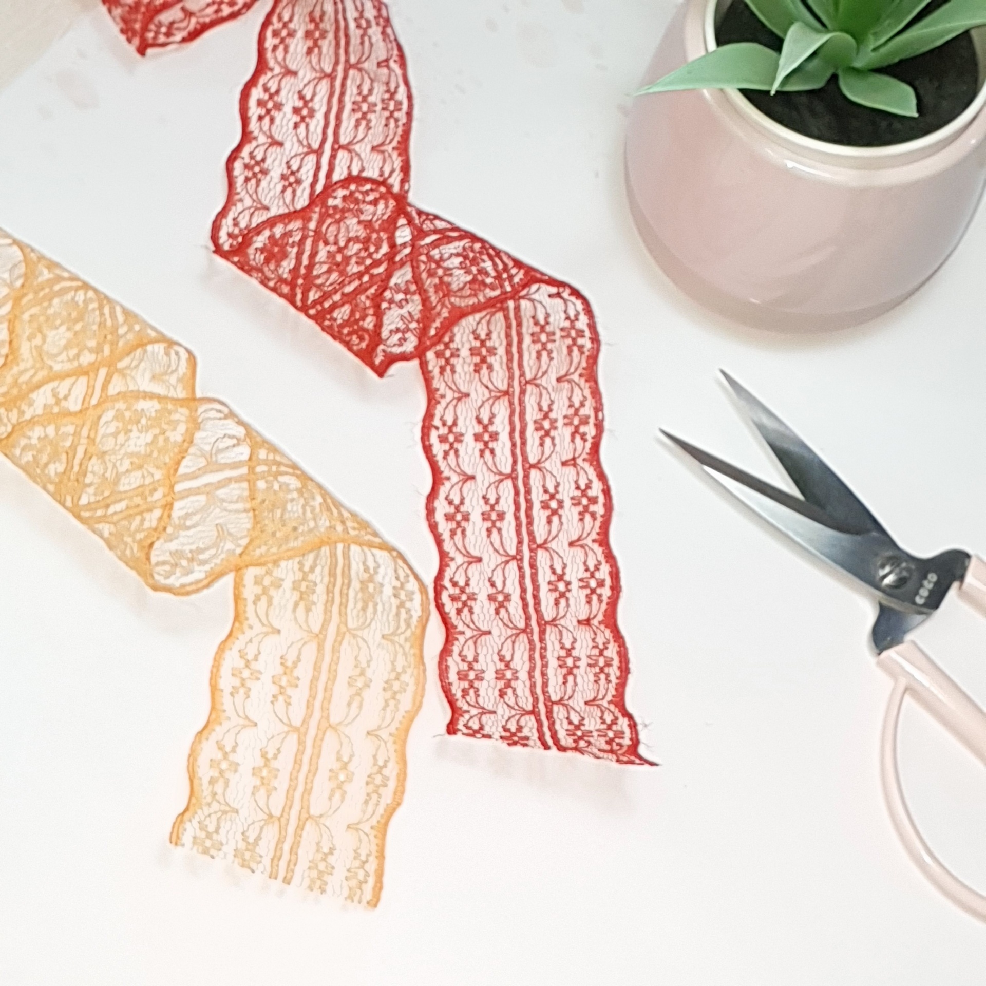 Tiras de renda vermelha e amarela, tesoura rosa e vaso com planta verde sobre fundo branco.