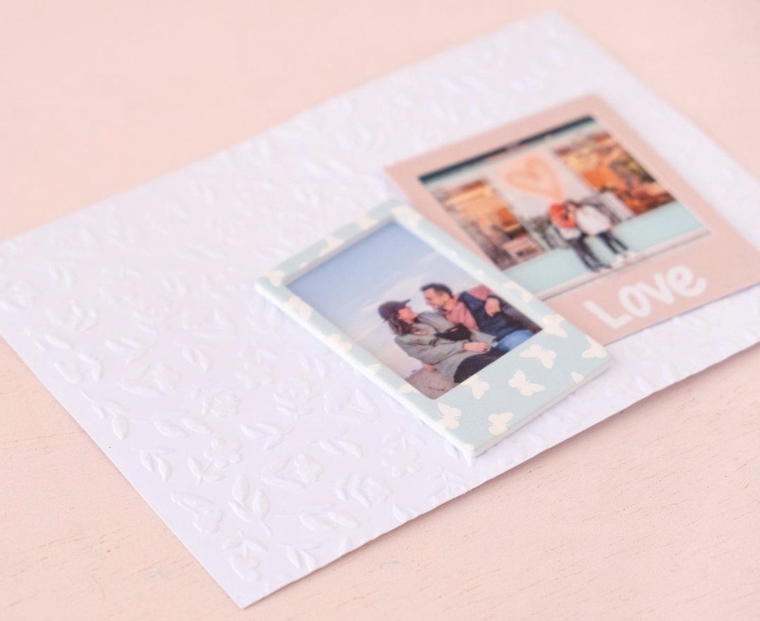 Dois porta-fotos tipo Polaroid, um azul com corações e outro rosa com a palavra 'Love' sobre fundo branco texturizado.