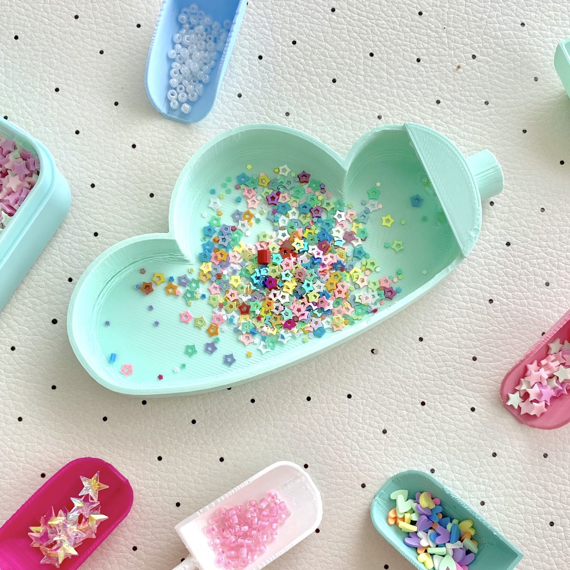 Bandejas coloridas com miçangas e ornamentos em superfície branca pontilhada.