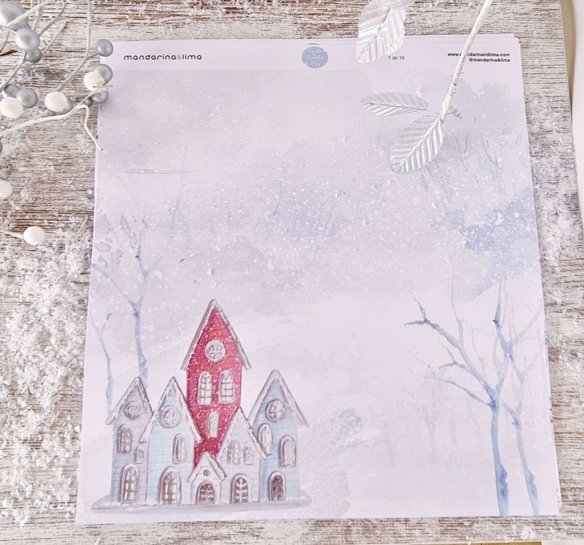 Bloco de papel decorativo com casas vermelhas e brancas, padrão de neve, sobre superfície de madeira clara com ramos prateados