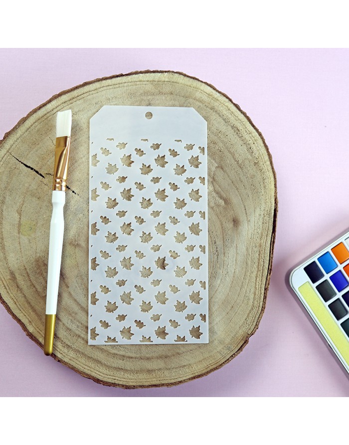 Stencil branco com padrão de folhas, pincel branco e dourado, tronco de madeira e caixa de tintas aquarela