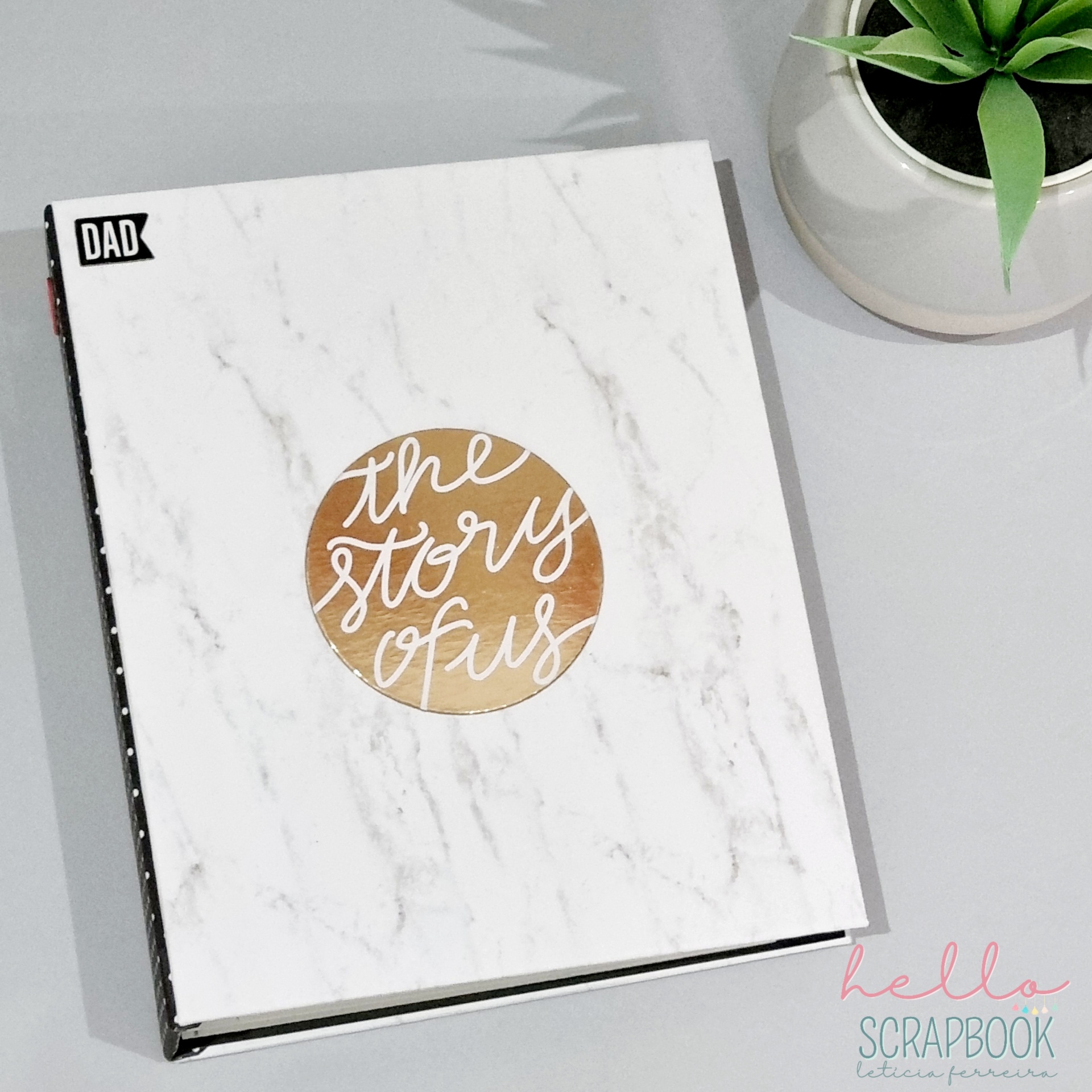 Livro com capa branca de efeito mármore, círculo dourado com texto 'the story of us' e etiqueta 'DAD' ao lado de planta em vaso branco