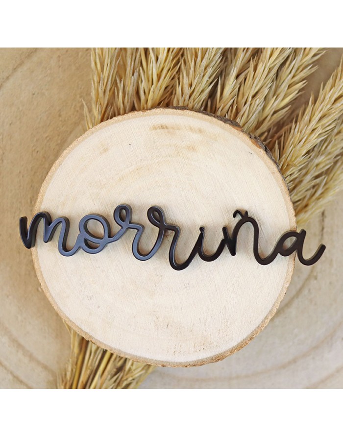 Produto decorativo em madeira preta com a palavra 'morriña' sobre base circular de madeira clara e plantas secas ao fundo.