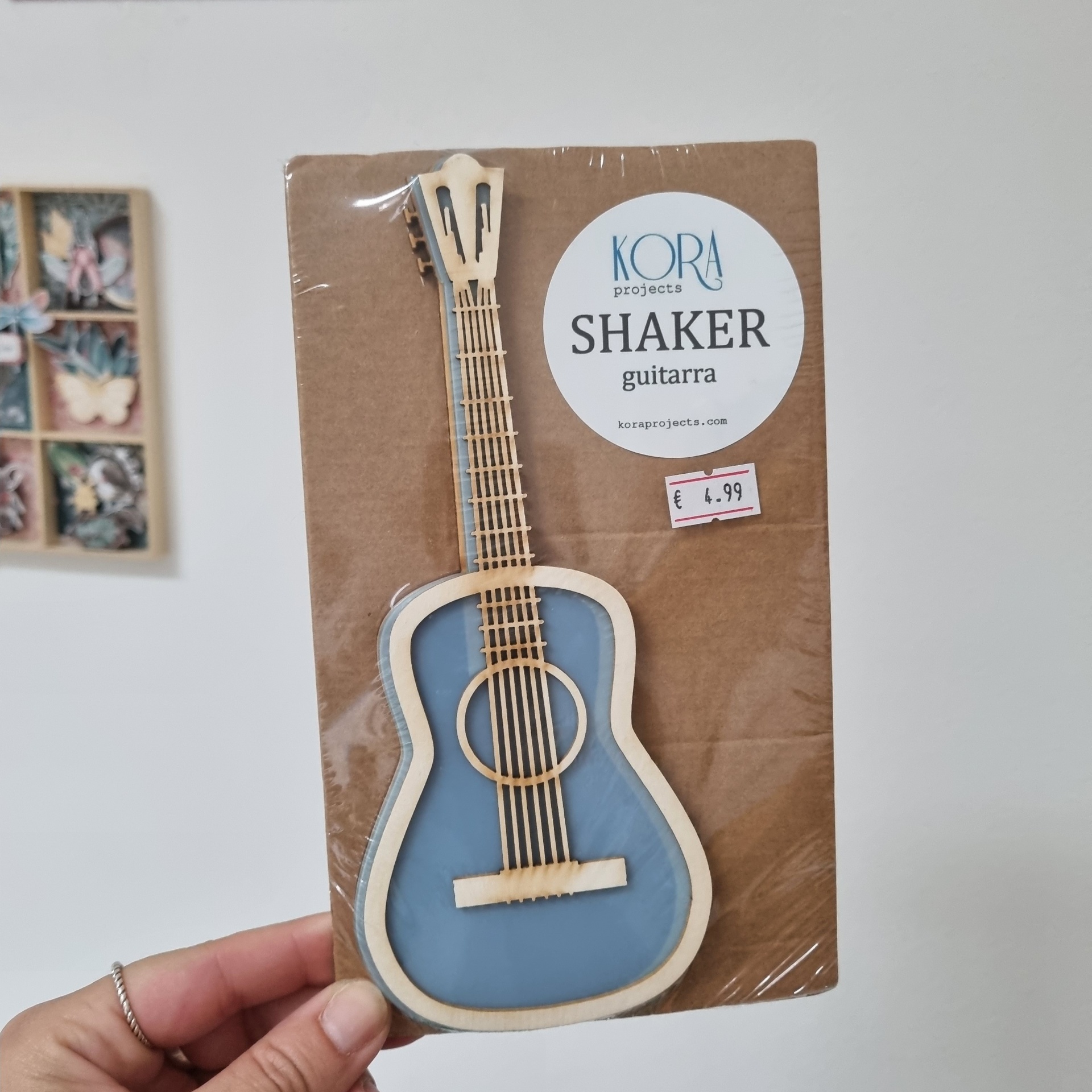 Guitarra artesanal em madeira clara e azul, embalado com etiqueta KORA projects SHAKER guitarra e preço de 4,99 euros