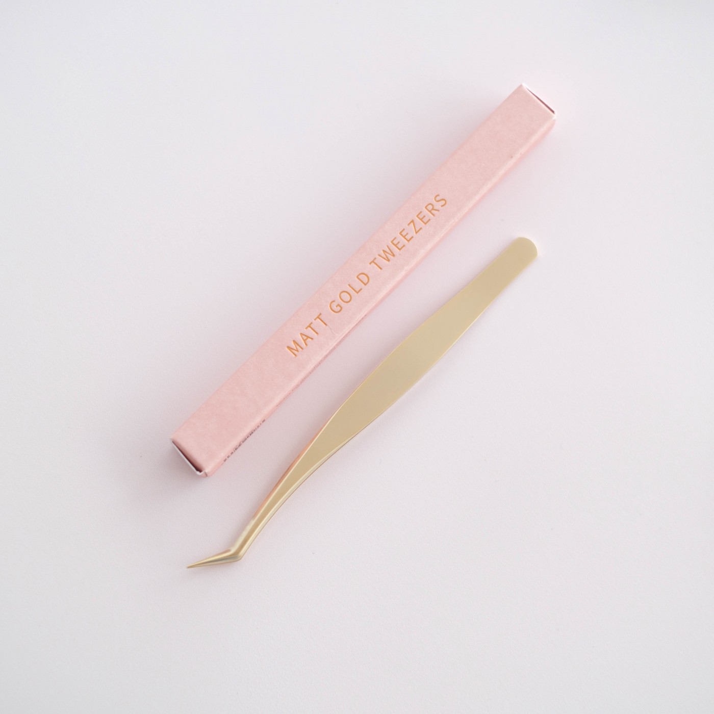 Pinça dourada mate com ponta inclinada e embalagem rosa com texto dourado