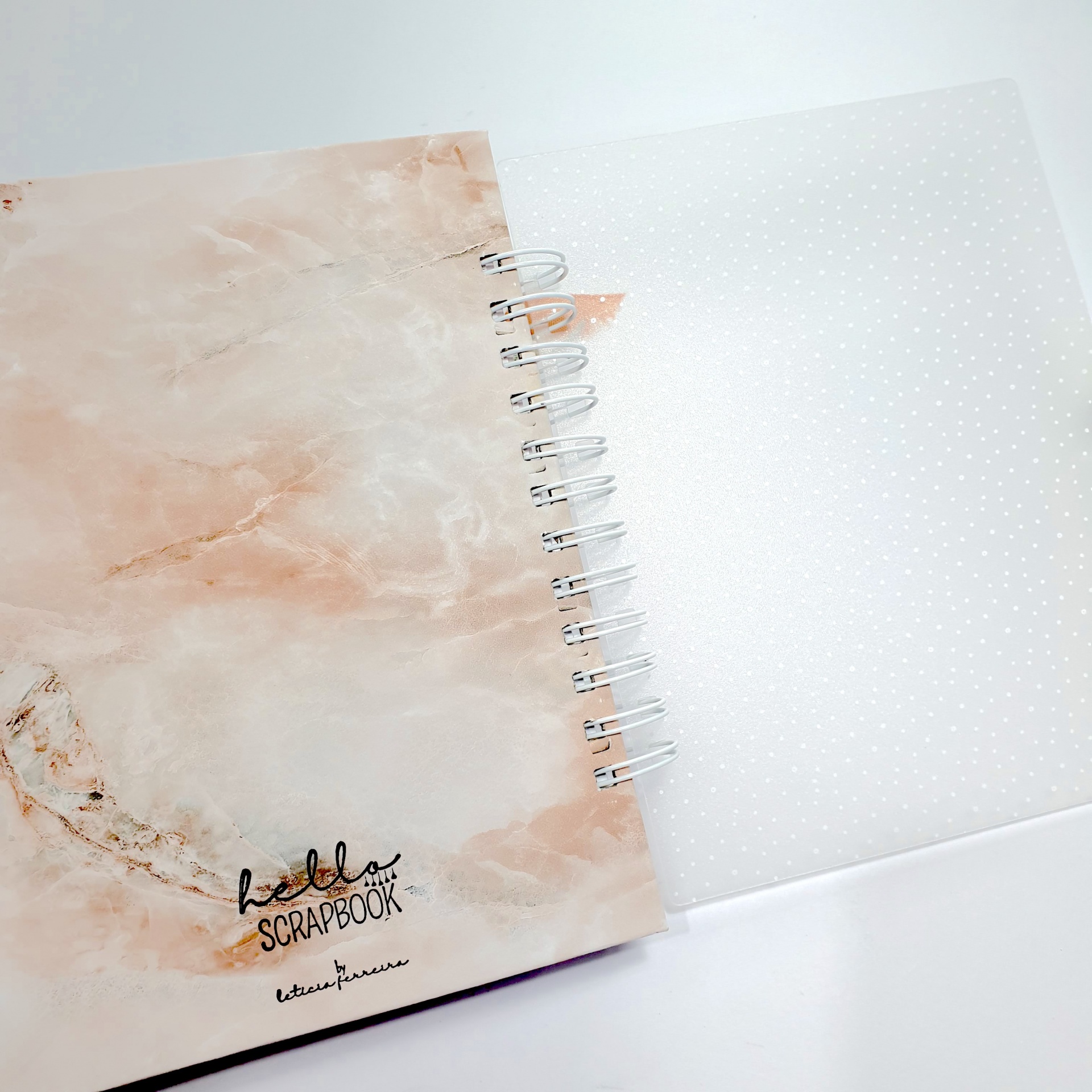 Caderno scrapbook com capa marmoreada bege e rosa, texto preto, encadernação branca, capa protetora transparente com pontos brancos