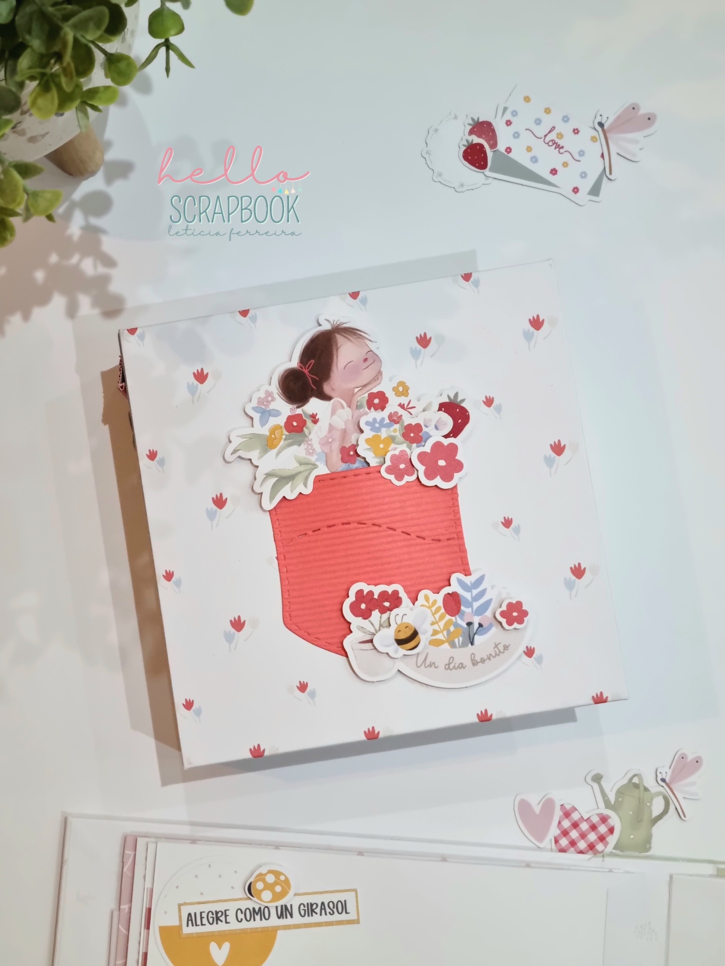 Caderno scrapbook com bolso vermelho e ilustração menina com flores, etiquetas adesivas e planta ao lado