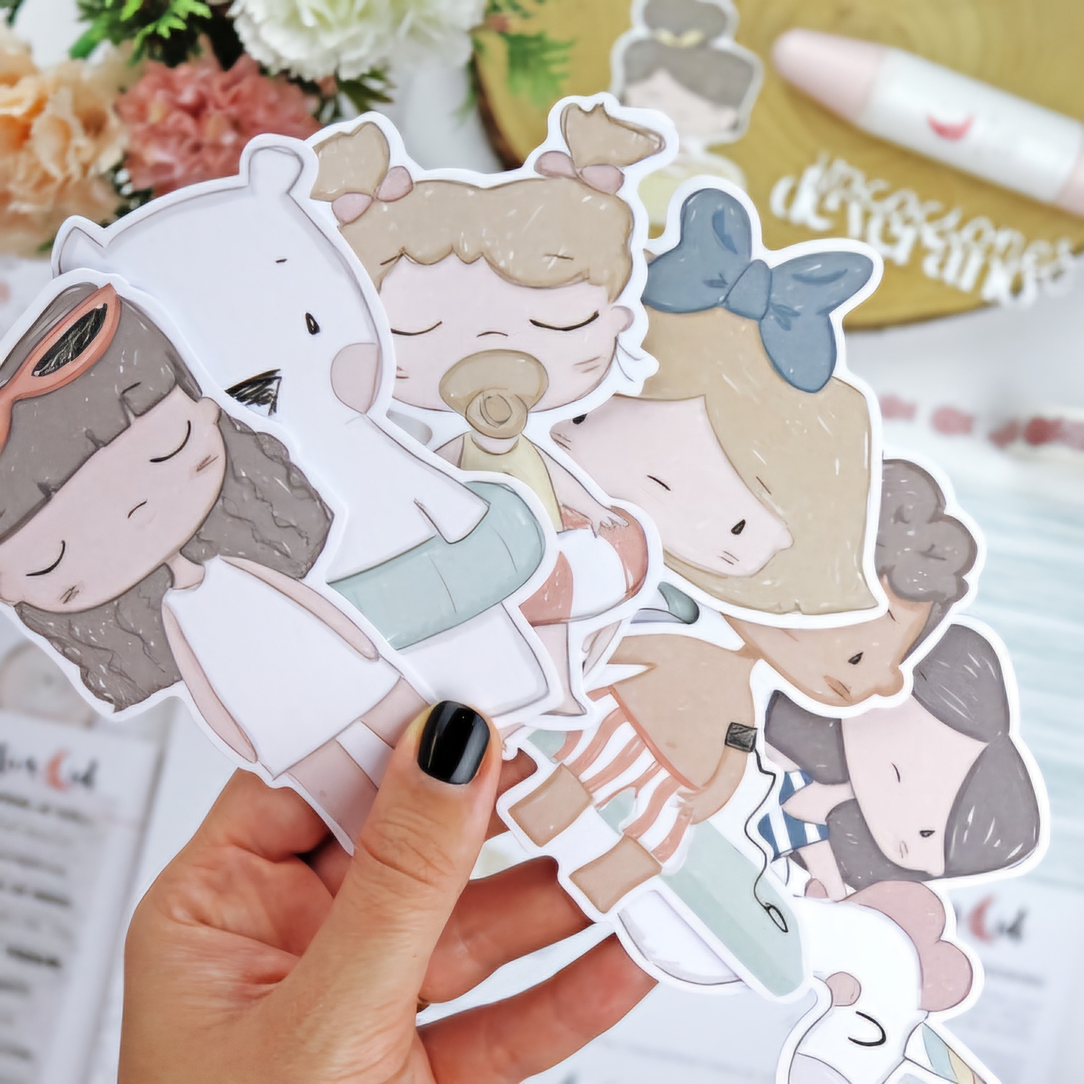 Autocolantes ilustrados de personagens femininas e urso branco segurados por mão com unhas pretas em fundo desfocado