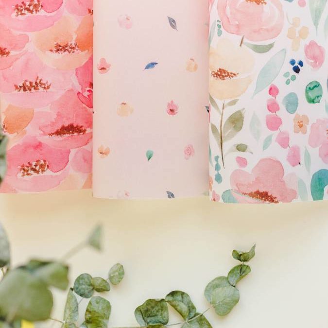 Rolos de papel com padrões florais coloridos em tons rosa e verde, sobre superfície clara com folhas verdes
