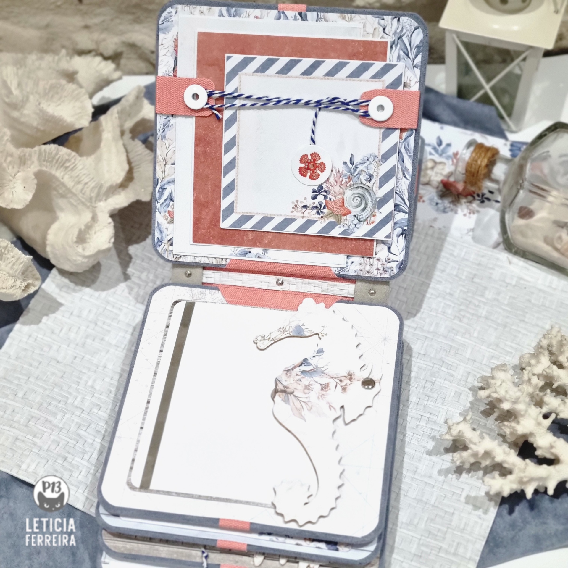 Álbum decorativo com capa em padrões marítimos azul, branco e vermelho sobre mesa com corais secos e lanterna branca