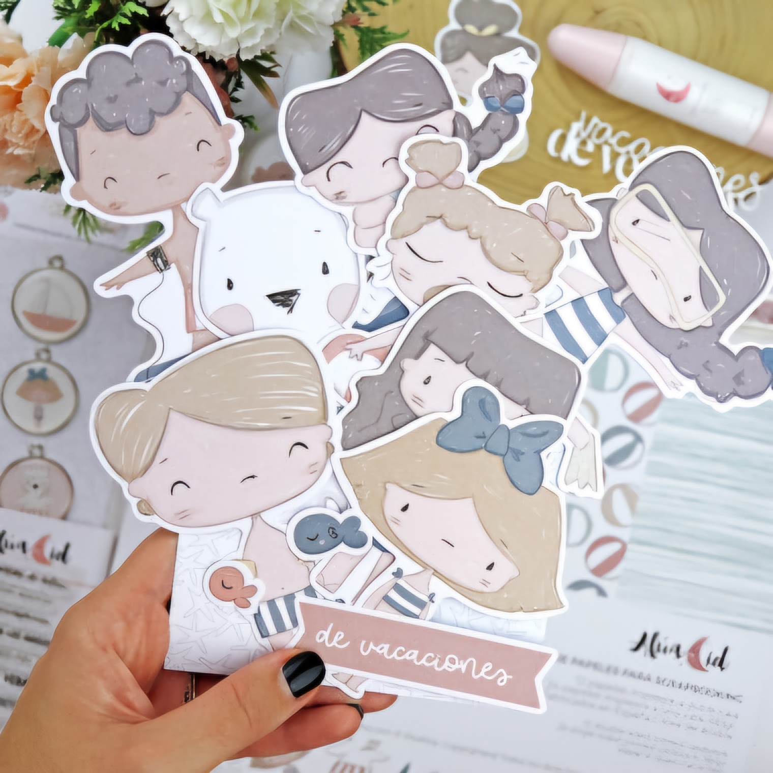 Cartões ilustrativos de personagens infantis coloridos segurados por uma mão, sobre mesa com flores e papelaria