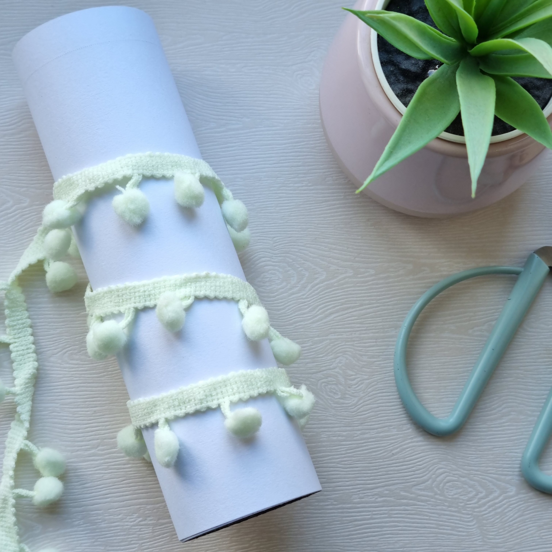 Rolo branco com fita de tecido creme com pompons ao lado de vaso rosa com planta e ferramenta azul